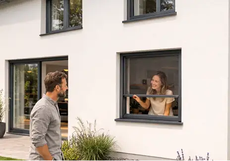 Frau öffnet ein Fliegengitter-Rollo am Fenster, während ein Mann davor steht