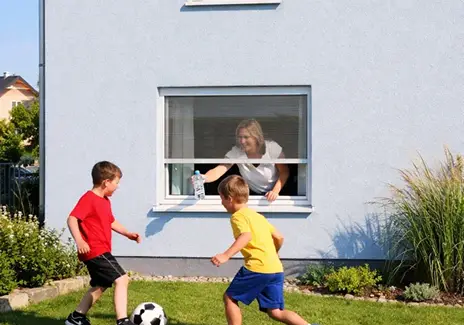 Frau öffnet ein Fliegengitter-Rollo am Fenster, während zwei Kinder im Garten Fußball spielen