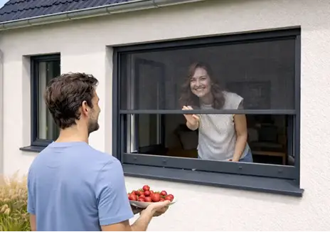 Frau öffnet ein Fliegengitter-Rollo am Fenster, während ein Mann draußen Tomaten hält
