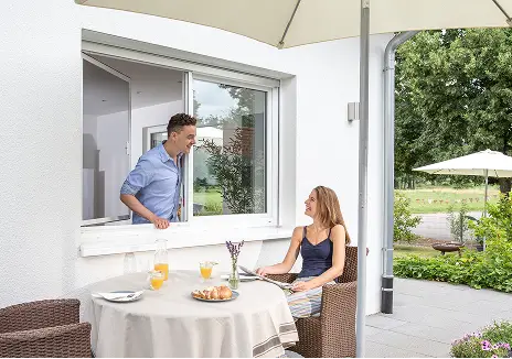 Mann steht an einem geöffneten Fenster mit Fliegengitter, während eine Frau auf der Terrasse sitzt